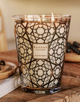 A lit scented candle in a clear glass jar with a black geometric Arabic pattern and gold trim, labeled 'Baabab Collection Arabian Nights', set on a rustic wooden table with decorative elements.