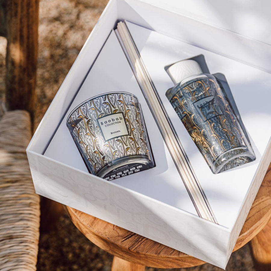 An art nouveau atmosphere is created on a wooden table, where a Baobab Brussels My First Baobab MFBBOXBRU scented candle and fragrance diffuser are neatly placed inside a delicate box.