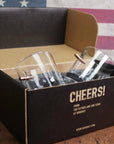 Clear glass bullet-shaped shot glasses in a black box with 'Cheers!' text against an American flag background.