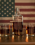 Clear whiskey glass decanter and two whiskey glasses with American flag design against an American flag background