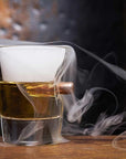 Glass with a bullet and liquid, surrounded by smoke on a wooden surface.