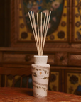 A reed diffuser bottle with a white textured cap and natural reed sticks, placed on a wooden table against a warm, patterned wooden background.