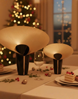 Dining table set for a festive meal with two modern ceramic vases, a glass, and small desserts in a softly lit room.