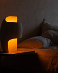 Modern table lamp with a warm glow on a bedside table in a dimly lit room.
