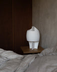 White sculptural table lamp on a wooden shelf next to a bed with gray bedding.