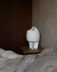 White sculptural table lamp on a wooden shelf next to a bed with gray bedding.