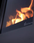 Close-up of a modern black wood burning fireplace or stove with visible orange flames through a glass window and the Dik Geurts logo on the front trim.