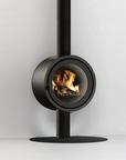 A modern black circular wood-burning fireplace with a visible flame, mounted on a vertical pipe and round base against a light gray wall.