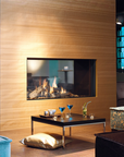 A modern linear gas fireplace with a black firebox insert set into a warm wood-paneled wall, styled in a contemporary lounge setting with a low black coffee table, blue cocktail glasses, and ambient lighting.