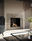 A modern gas fireplace with a white and gray marble-look surround, lit with visible flames, set in a styled living room with a blue armchair, vase of flowers, and glass coffee table.