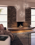 A modern living room featuring a stone fireplace with a gas fireplace, large black-framed windows, a glass bubble pendant light, a beige sofa with a brown pillow and blanket, and a glass coffee table.