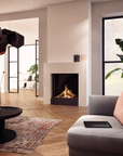 A modern living room interior featuring a white fireplace with a gas lit fireplace, a gray upholstered armchair with a pink cushion, a black round coffee table, wood flooring, and large windows.