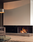 A modern linear gas fireplace with a visible fire burning inside, set into a low stone hearth in a contemporary living room. A gray upholstered armchair, a floor lamp, and a glass coffee table are visible in the foreground.