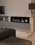 A modern linear gas fireplace with glowing yellow-orange flames, set into a light beige wall above a gray concrete mantel in a contemporary living room. A black floor lamp with a drum shade stands to the right, and a dark textured ottoman is in the foreground.