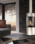 A modern cylindrical wood-burning fireplace with a glass front and visible flame, set in a contemporary living room with a dark stone wall, gray sofa, and large windows.