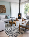 A modern living room featuring a white sectional sofa, two wooden-framed armchairs with gray cushions, a round wooden coffee table, a black wood-burning fireplace, and a large black-and-white geometric patterned area rug.