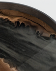 Close-up of a dark ceramic plate or bowl showing a glossy black interior with a textured, organic brown and bronze marbled pattern along the rim against a light gray background.
