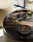 A round black glass coffee table with a reflective dark surface, styled with two textured clear drinking glasses, a tall amber glass bottle, and a small dark plate with nuts. Part of a beige woven pouf is visible on the left, and a window with outdoor light appears in the background.