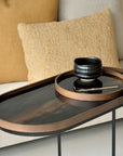 An oval side table with a glossy black glass top and warm wood-toned rim, styled with a small black bowl and spoon on a matching tray, set in a living room beside beige and mustard pillows.