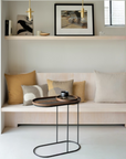 A modern interior scene with a light wood bench seat styled with beige and mustard pillows, a small oval side table in front, wall shelves, pendant lights, and framed art in a bright neutral room.