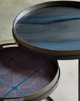 Two round nesting side tables with dark metal frames and trays. The top table has a dark blue-gray marbled glass top, and the bottom table has a dark purple geometric patterned tray.