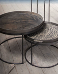 Two round nesting side tables with dark metal frames and textured tabletops, one featuring a dark wood grain pattern and the other a circular dot design, set on a rustic wooden floor.
