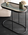 An oval-shaped side table with a dark glass top and a black metal frame base, styled with a small bowl and a vase of dried flowers on a dark wood floor next to a light beige sofa.