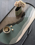 An oval glass-top coffee table with a black metal frame and curved legs, styled with a ceramic vase of flowers, a small bowl, and a few sugar cubes on a dark wood floor.