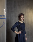 Woman in a dark blue dress standing between two modern chandeliers against a gray wall.