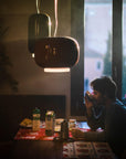 Person sitting at a table in a dimly lit room with a window in the background and a round and long suspension light from Foscarini hanging from the ceiling.