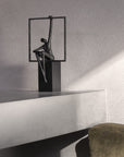 Sculpture of a person sitting on a square platform against a gray wall.