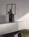 Sculpture of a person sitting on a square platform against a gray wall.