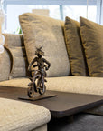 Bronze sculpture of a person on a bike on a coffee table in front of a beige sofa with green pillows.