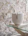 White ceramic sculpture on a glass table against a textured stone wall