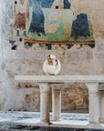 White ceramic sculpture on a pedestal in front of a mural on a stone wall.