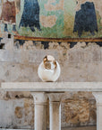 White ceramic sculpture on a pedestal in front of a mural on a stone wall.