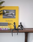Book titled 'The Impossible Collection of Design' by Assouline on a shelf with decorative bronze statues and plants.