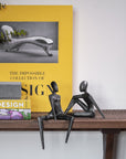 Book titled 'The Impossible Collection of Design' by Assouline on a shelf with decorative bronze statues and plants.