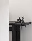 Two black sculptures of people sitting on a ledge against a white wall.