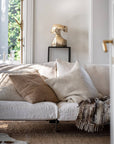 White sofa with beige and brown cushions in a bright room with natural light and a polished bronze sculpture on a high pedestal.