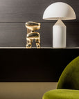 White lamp and polished bronze sculpture on a dark surface with a green chair in the foreground.