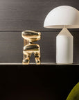 White lamp and polished bronze sculpture on a dark surface with a green chair in the foreground.