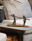 Bronze sculpture of two figures carrying a log on a wooden base, placed on a coffee table.
