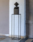 Black geometric bronze sculpture on a tall black stand against a white brick wall.