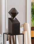 Abstract black metal sculpture on a stand against a neutral wall with a plant in the background