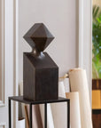 Abstract black metal sculpture on a stand against a neutral wall with a plant in the background