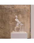 White sculpture of a seated figure on a pedestal against a textured stone wall.