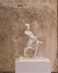 White sculpture of a seated figure on a pedestal against a textured stone wall.