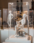 Sculpture of a seated figure with a glass ball inside, displayed in a clear case.