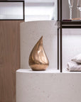 Bronze teardrop sculpture on a white surface with a blurred background